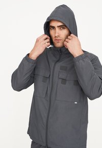 Graue Kapuzenjacke mit einem Frontreißverschluss, elastischen Bündchen und zwei Klappentaschen. Das Material wirkt leicht und glatt.