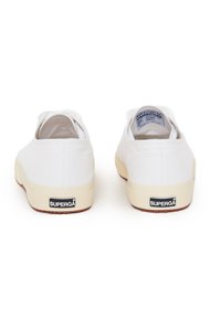 Sneakers bianchi in tela con suola in gomma beige, caratterizzate da un motivo testurizzato, logo sul retro e design minimalista.