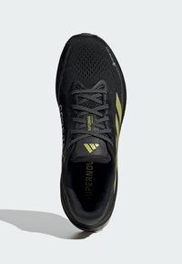 Black running shoe with mesh upper, green accents, and flat laces. Features textured sole and embossed brand labeling on the side and tongue.