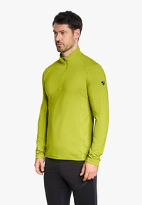 Grünes Langarmshirt mit hohem Kragen, leichtem Stoff und einem Viertelreißverschluss. Es hat ein kleines schwarzes Logo auf dem linken Ärmel.