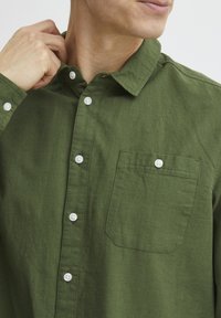 Camisa de algodón verde con cuello abotonado, botones blancos y un solo bolsillo en el pecho con un acabado limpio y texturizado.