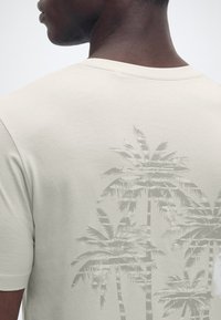 Άνδρας που φοράει ανοιχτό μπεζ μπλουζάκι με διακριτικό γραφικό σχέδιο φοίνικα στην πλάτη, απεικονιζόμενος από τον ώμο έως τον λαιμό με φόντο απλό.