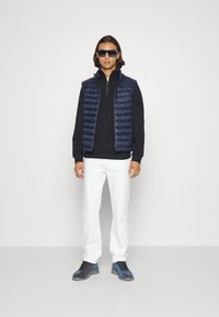 Vesta puffer navy peste un top cu guler cu fermoar negru, pantaloni albi tip tapered și încălțăminte albastră. Modelul poartă ochelari de soare închiși. Fundal simplu.