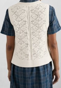 Kremowa dzianinowa kamizelka z geometrycznym wzorem koronki, teksturowaną górą i jednolitym ściągaczem, noszona na niebieską sukienkę koszulową w kratę.