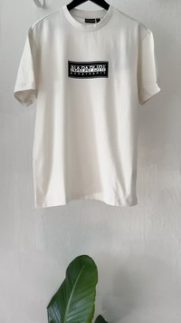 Camiseta de algodón color crema con mangas cortas, que presenta un logo rectangular negro con el texto "NAPAPIJRI geographic" centrado en la parte frontal.