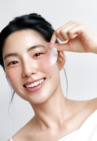 Patches hydrogel pour les yeux à finition nacrée, appliqués sous les yeux, portés par une femme souriante aux cheveux foncés, vêtue d'un haut blanc.