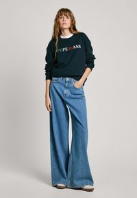 Marinblå sweatshirt med "Pepe Jeans"-logotyp i rött och grönt, kombinerad med vida, ljusblå randiga denimjeans. Avslappnade skor.