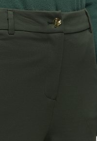 Pantalon vert foncé avec un bouton abstrait doré et des passants de ceinture, porté avec un haut vert texturé.