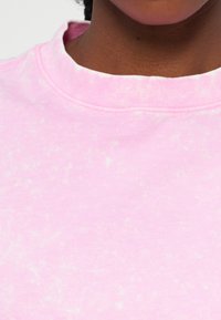 Κοντινό πλάνο του λαιμού και του άνω μέρους του στήθους που φοράει ροζ, ελαφρώς μαρμαρωτό φούτερ με στρογγυλή λαιμόκοψη σε σκούρο τόνο επιδερμίδας.