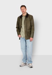 Brown leather jacket with a dark collar, green shirt, light blue jeans, and gray sneakers. The jacket features snap buttons and side pockets.