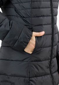 Zwarte pufferjack met horizontale naden, een volledige ritssluiting en een hand in een zijzak. Materiaal lijkt glanzend en glad te zijn.