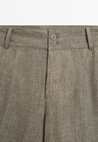 Gros plan de la taille d’un pantalon en lin beige avec deux boutons et des passants de ceinture, montrant la texture et les détails des coutures.