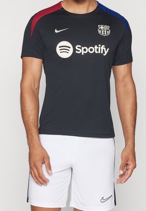 Homme portant un maillot de football sombre FC Barcelone Nike avec le logo Spotify et un short Nike blanc sur fond uni.