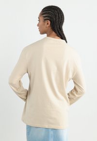 Beige long-sleeve shirt made of soft cotton, featuring a relaxed fit. The back has a round neckline and minimal seam detailing.