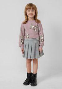 Ein junges Mädchen mit hellbraunem Haar, das einen lila Pullover mit Teddybären, einen grauen Faltenrock und schwarze Schnürboots trägt, steht vor einer weißen Wand.