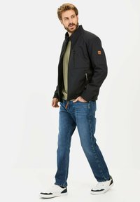 Sort softshell jakke med høj krave, lynlåslommer og en tan logo-lap, båret over en grøn skjorte, parret med blå jeans og hvide sneakers.
