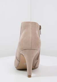 Botín de ante beige con tacón alto, que cuenta con un zipper lateral y un detalle de costura visible en la parte trasera. Textura suave.