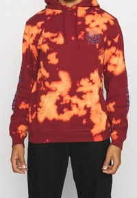 Felpa con cappuccio bordeaux con pattern tie-dye arancione, tasca frontale a marsupio, cappuccio regolabile con cordino e dettagli testuali blu sulle maniche.