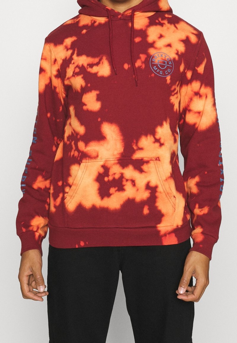 Sudadera color granate con patrón de tie-dye naranja, bolsillo frontal tipo canguro, capucha ajustable con cordón, y acentos de texto azul en las mangas.