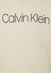 Sudadera beige con un gran logo negro de "Calvin Klein" en el pecho; hecha de material suave y con un bolsillo tipo canguro en la parte frontal.