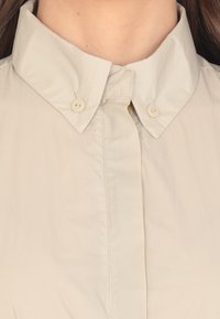 Camicia beige con colletto, dotata di una patta con bottoni, texture liscia e una leggera lucentezza. Due bottoni visibili sul colletto per la chiusura.