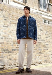 Modrý vzorovaný sakový kabát z látky s geometrickými vzormi, prekrývajúci hnedú košeľu. Krémové nohavice a tmavohnedé topánky dopĺňajú celkový vzhľad.