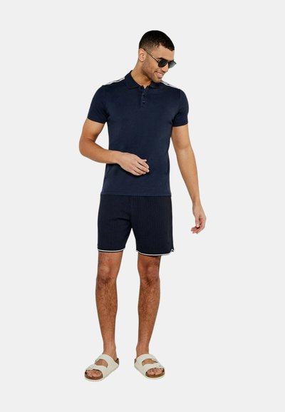 Polo shirt navy con tre bottoni, dettagli a strisce in contrasto sulle spalle, abbinato a pantaloni corti testurizzati scuri e sandali beige.