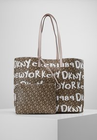 Sac fourre-tout DKNY marron et beige avec des poignées rose clair, motif texte blanc "DKNY New York 1989" et petite pochette assortie sur un socle blanc.