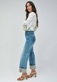Blusa floral blanca con mangas abullonadas, combinada con jeans de tiro alto en un azul claro con dobladillo enrollado y sandalias de tacón cuadrado beige.