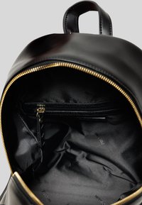 Interno di uno zaino in pelle nera con texture liscia. Presenta una cerniera dorata, fodera nera e una tasca con cerniera per l'organizzazione.