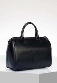 Sac à main en cuir noir avec deux anses et une fermeture éclair sur le dessus, posé sur un piédestal hexagonal blanc devant un fond uni.