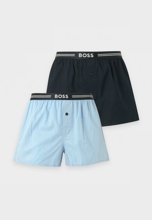 Muskelbepackter, oberkörperfreier Mann steht barfuß und trägt hellblaue Boxershorts mit schwarzem Bund mit der Aufschrift „BOSS“ vor einem einfarbigen weißen Hintergrund.