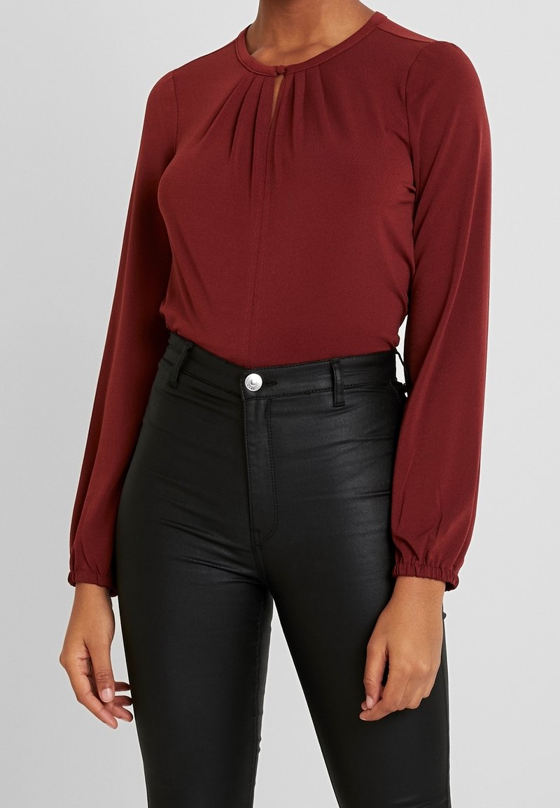 Burgundowa bluzka z długim rękawem i okrągłym dekoltem, z plisowanym przodem oraz wycięciem w kształcie łezki, w zestawieniu z wysokimi czarnymi spodniami z ekologicznej skóry.
