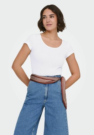 Vrouw met kort bruin haar, een wit geribbeld kortärmelig shirt, blauw hooggetailleerd spijkerbroek en een bruine sjaal met patroon als riem gestrikt rond de taille.