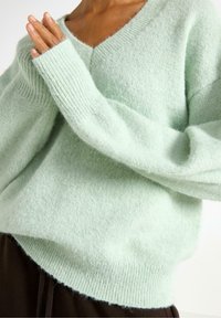 Vaaleanvihreä ylisuuri neulepaita, joka on valmistettu pehmeästä ja pörröisestä materiaalista. Siinä on V-pääntie, leveät ribatut hihansuut ja rento istuvuus.