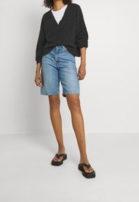 Cárdigan gris oscuro abotonado sobre una camiseta blanca, combinado con pantalones cortos de mezclilla azul claro. El conjunto se completa con chanclas negras de goma gruesa.