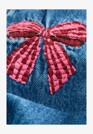 Tikkitud denimpind, millel on suur roosa pael keerukate kootud detailidega. Taust on tavaline sinine denimtöötlus.