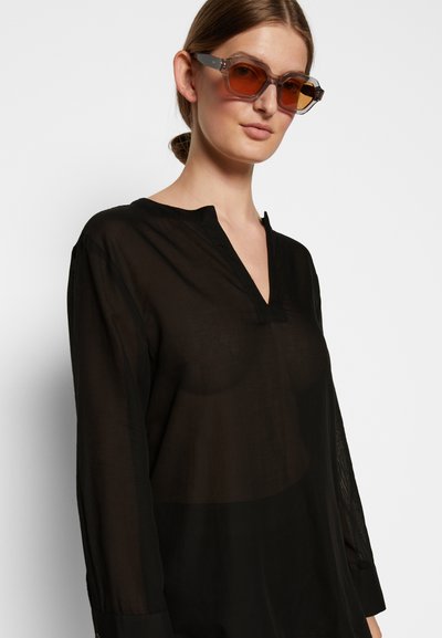 Camisa preta de manga longa feita de material transparente, com decote em V. Acompanhada de óculos de sol angulares, com lentes castanhas e armação translúcida.