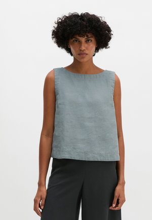 Femme aux cheveux bouclés portant un haut bleu clair sans manches et un pantalon noir, debout devant un fond blanc uni.