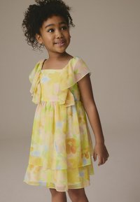 Robe florale jaune avec des volants superposés, décolleté carré et manches courtes. Fabriquée dans un tissu léger et semi-transparent avec des accents pastel.
