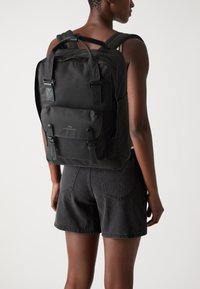 Sac à dos en tissu noir avec des bretelles rembourrées ajustables, une poignée supérieure, une poche avant et des accents en boucle. Texture lisse et design minimaliste.