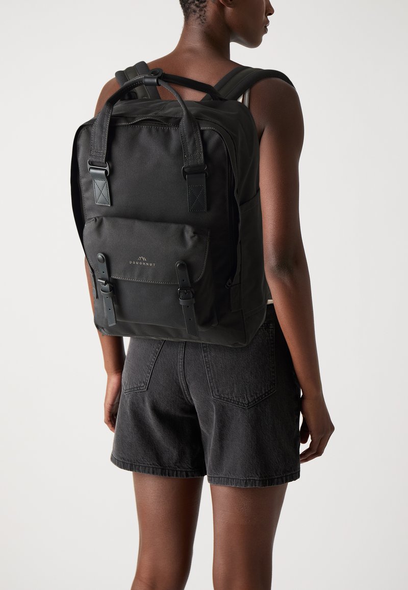 Sac à dos en tissu noir avec des bretelles rembourrées ajustables, une poignée supérieure, une poche avant et des accents en boucle. Texture lisse et design minimaliste.
