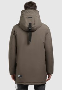 Rudas izoliuotas parkas su aukštu apykaklė ir reguliuojama kepure, su sagtimi ir etikete kairiajame apatinio šone.