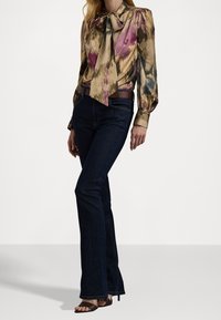 Žena v vzorované saténové blůze s kravatou, tmavých bootcut džínách s hnědým opaskem a hnědých sandálech na podpatku, stojící na bílém pozadí.