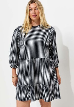 Kobieta w czarno-białej sukience w kratkę gingham z długimi bufiastymi rękawami i warstwową spódnicą stoi na jednolitym tle.