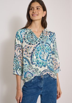 Femme portant un chemisier à motif floral bleu et rose avec un col à nouer et des manches trois-quarts, assorti à un jean bleu.