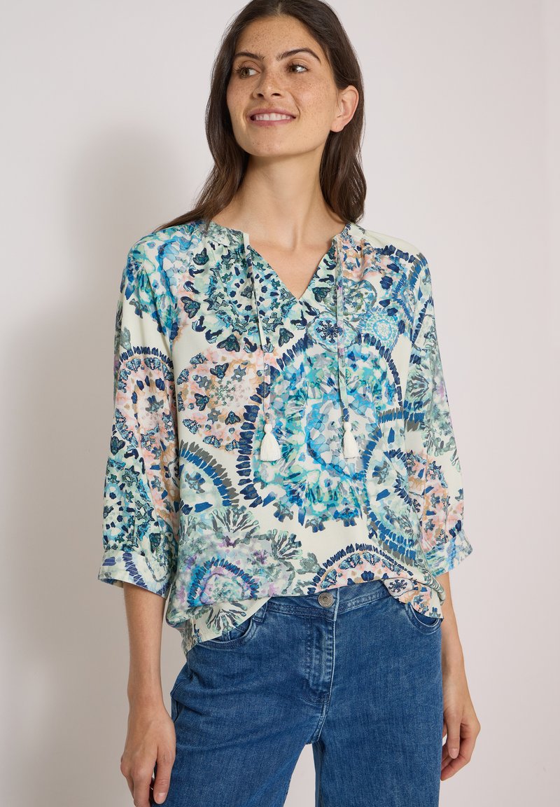 Femme portant un chemisier à motif floral bleu et rose avec un col à nouer et des manches trois-quarts, assorti à un jean bleu.