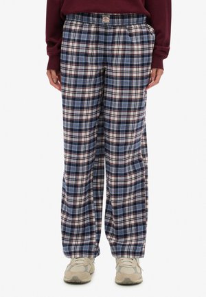 Personne portant un pantalon à carreaux bleu, blanc et rouge à coupe ample, associé à des baskets beige et un haut à manches longues bordeaux.