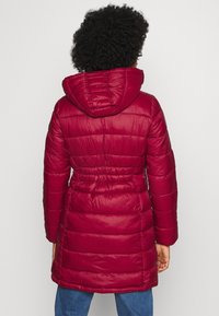 Mujer vista desde atrás con un abrigo acolchado con capucha rojo, combinado con jeans azul, contra un fondo liso.