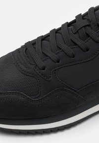 Black athletic shoe featuring a combination of textured mesh and smooth suede materials. Lace-up design with a white rubber sole and subtle stitching.